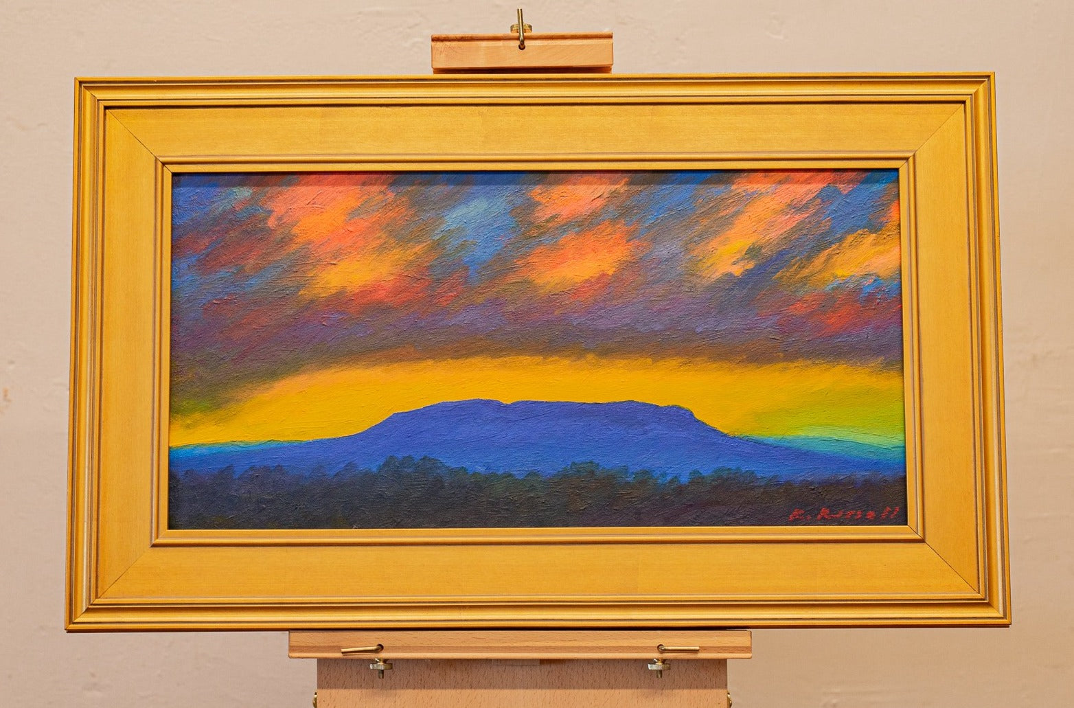 Blue Mountain by Ryan Russell. In a gold frame, resting on an artist's easel. Featuring a blue-toned House Mountain under a colorful evening sky.